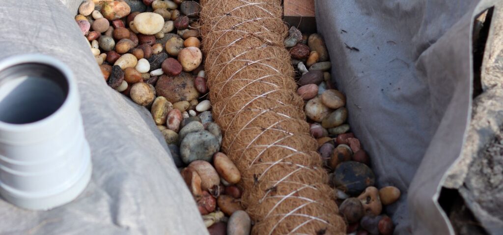 Exterior French drain trench dug for crawl space waterproofing in Raleigh, NC, with gravel packed around a fabric-wrapped drain pipe.