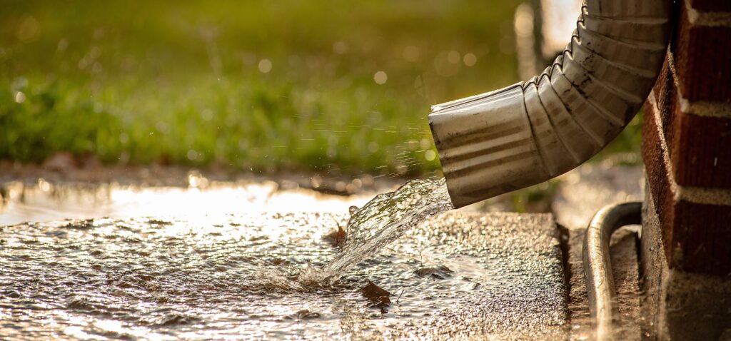 Rainwater flows from a gutter downspout onto the ground near a home’s foundation, showing runoff that can lead to basement flooding if not properly managed.