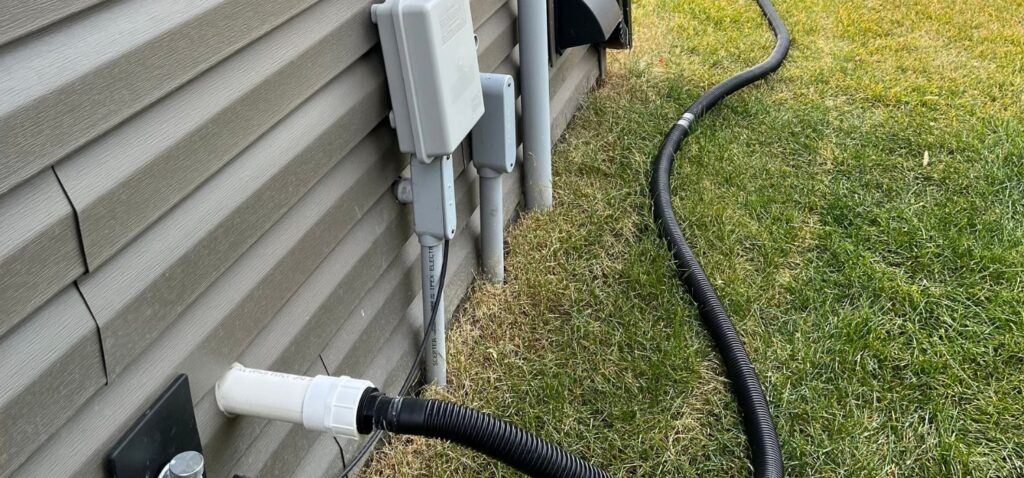 A black corrugated drain hose connected to a sump pump discharge pipe carries water away from the exterior wall of a home to prevent basement flooding.